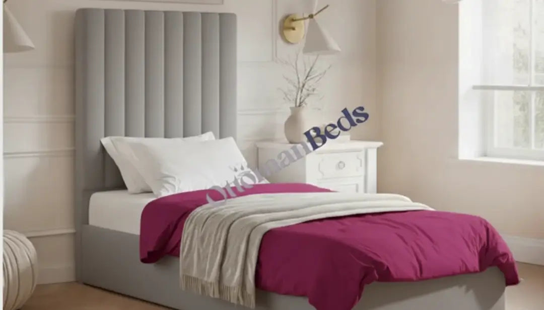 A grey Single Ottomans bed with a tall padded headboard, white pillows, and a purple duvet, styled in a bright bedroom with a bedside table and window.